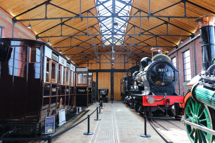 Vintage steam locomotives displayed indoors with a wooden roof and brick walls.