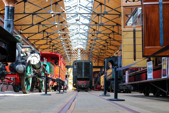 Vintage trains inside a railway museum with wooden and steel beams overhead.