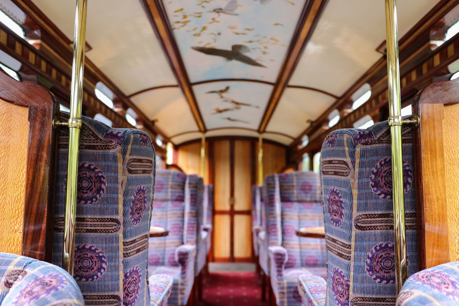 Vintage train car interior with patterned seats and bird-themed ceiling artwork.