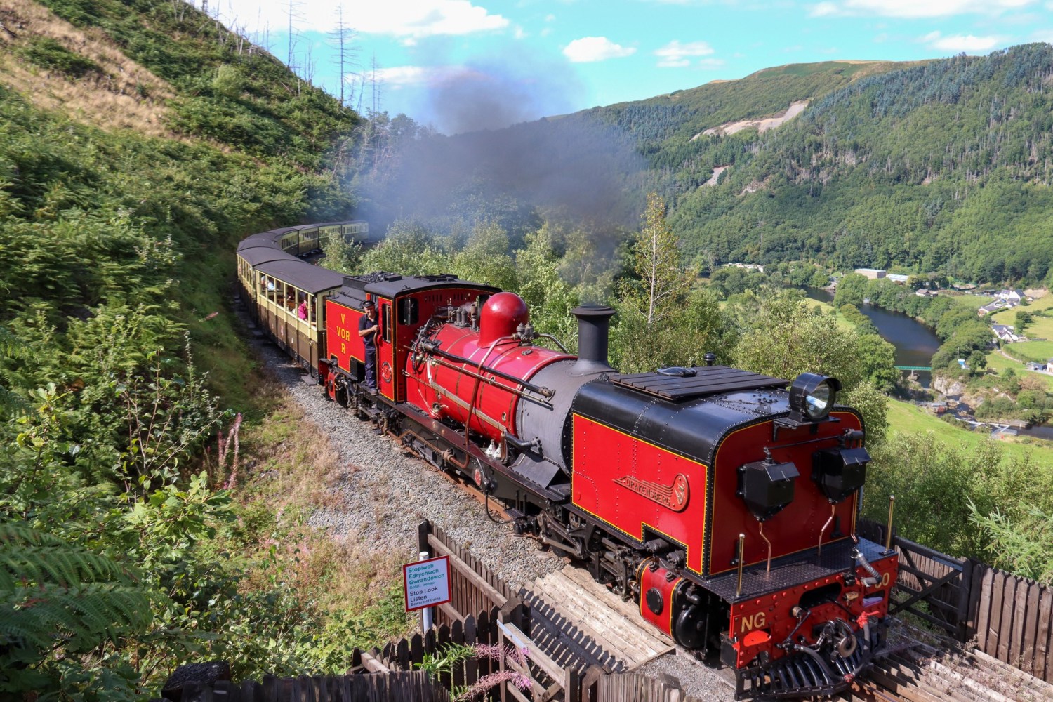 Red steam train on a scenic hillside track with mountains and greenery.