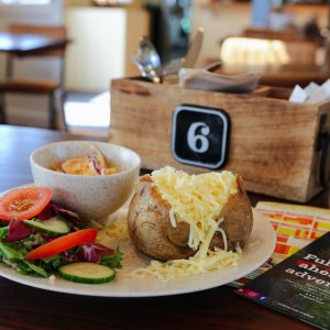 Plate with salad, baked potato, and cheese beside a bowl and wooden box on a table with flyers.