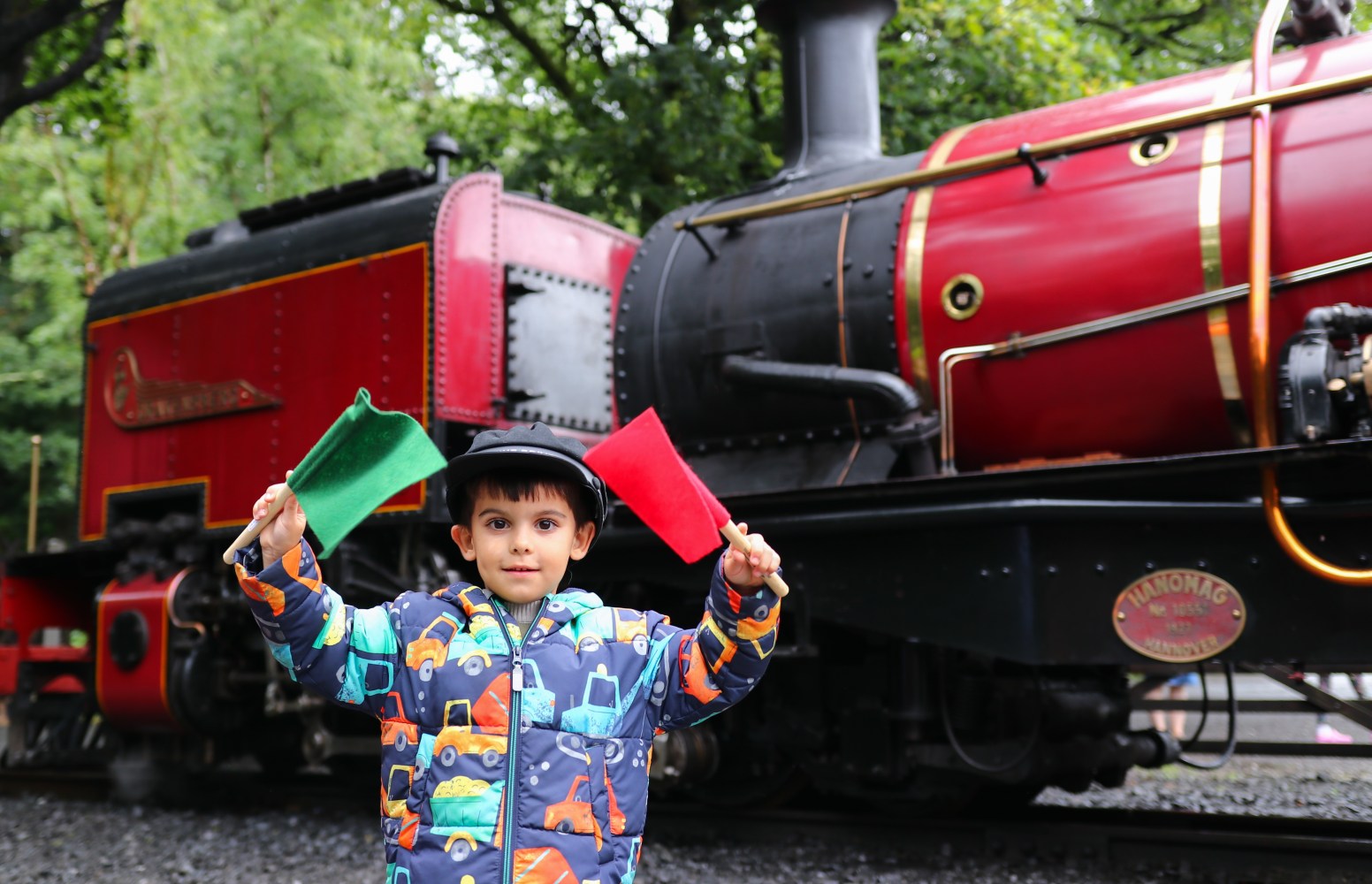 Child holding red and green flags in front of a vintage red steam train.