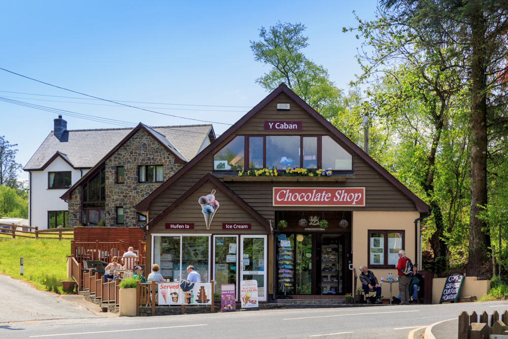 Chocolate shop with outdoor seating and adjacent house, surrounded by trees and a clear sky.