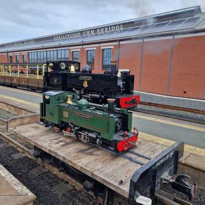 Miniature steam locomotives on a flatbed at a train station with a brick building in the background.