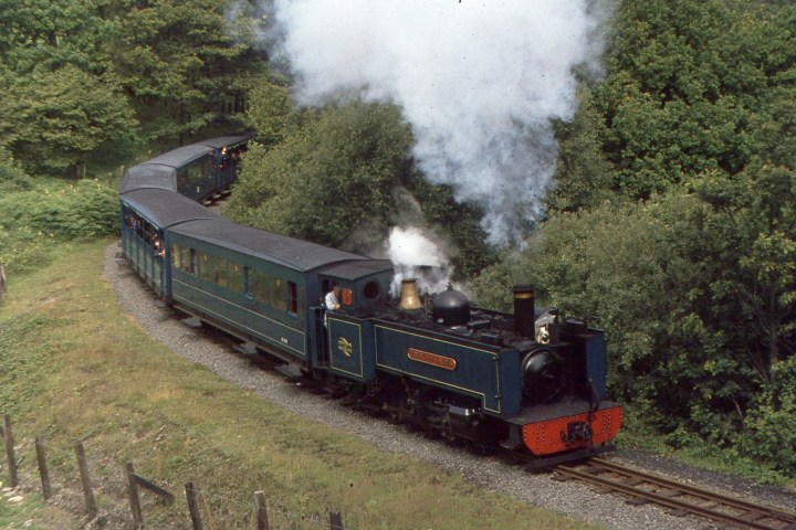 Steam train with blue carriages traveling through a wooded area.