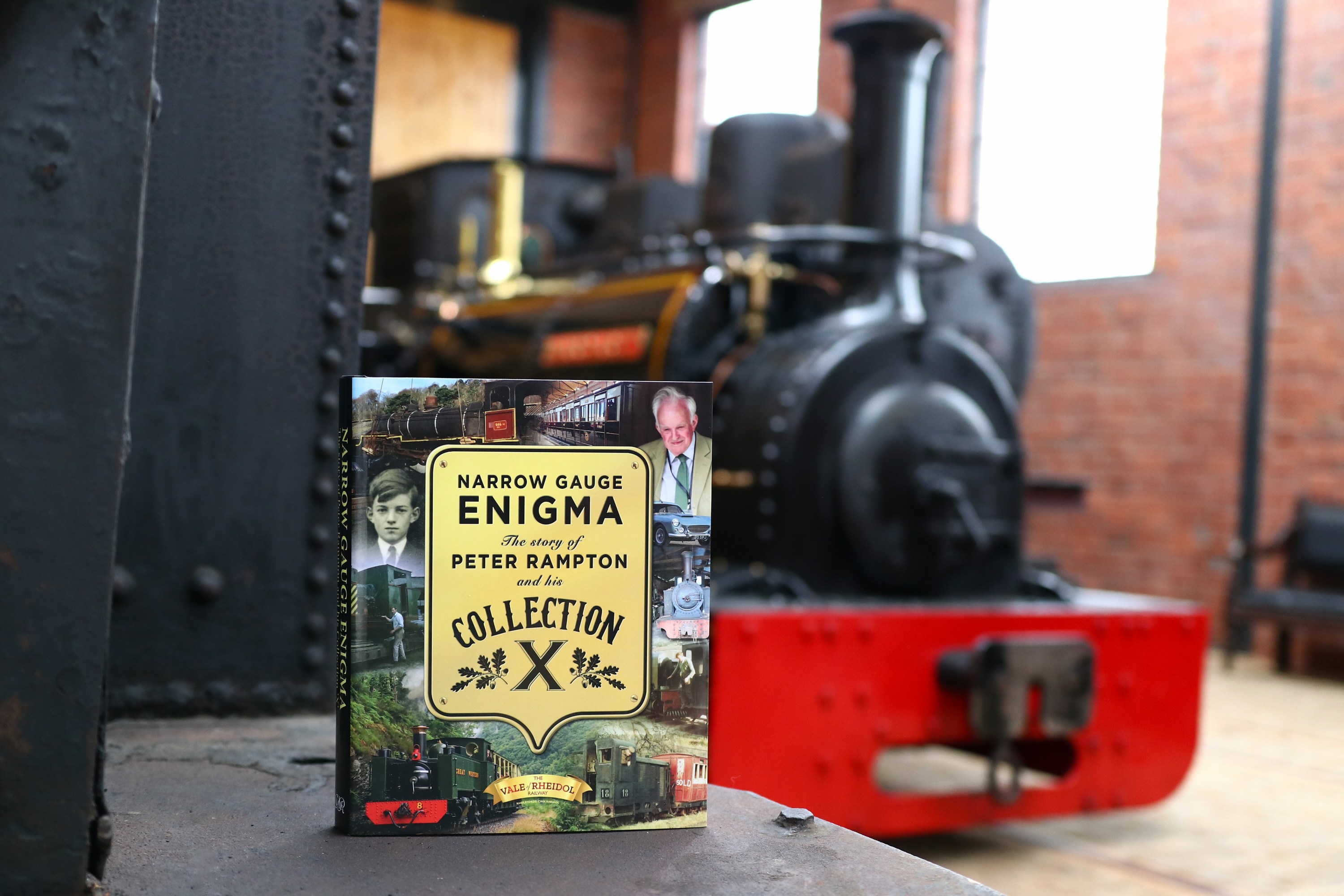 Book titled 'Narrow Gauge Enigma' in front of a vintage train inside a brick-walled room.