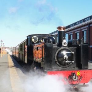 Smiling train conductor in window with vintage steam train at platform on a sunny day.