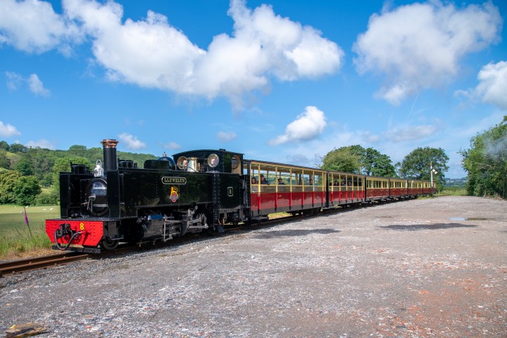 Steam train with black engine and red carriages on a sunny day, countryside in the background.