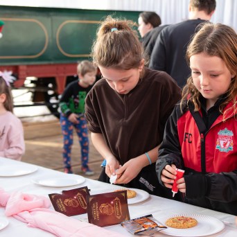 Children decorate cookies at a table, wearing casual clothes and holiday-themed attire.