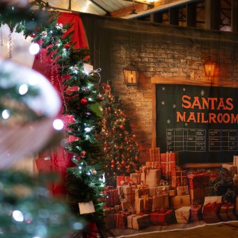 Festive room with decorated Christmas trees and a 'Santas Mailroom' sign, surrounded by presents.
