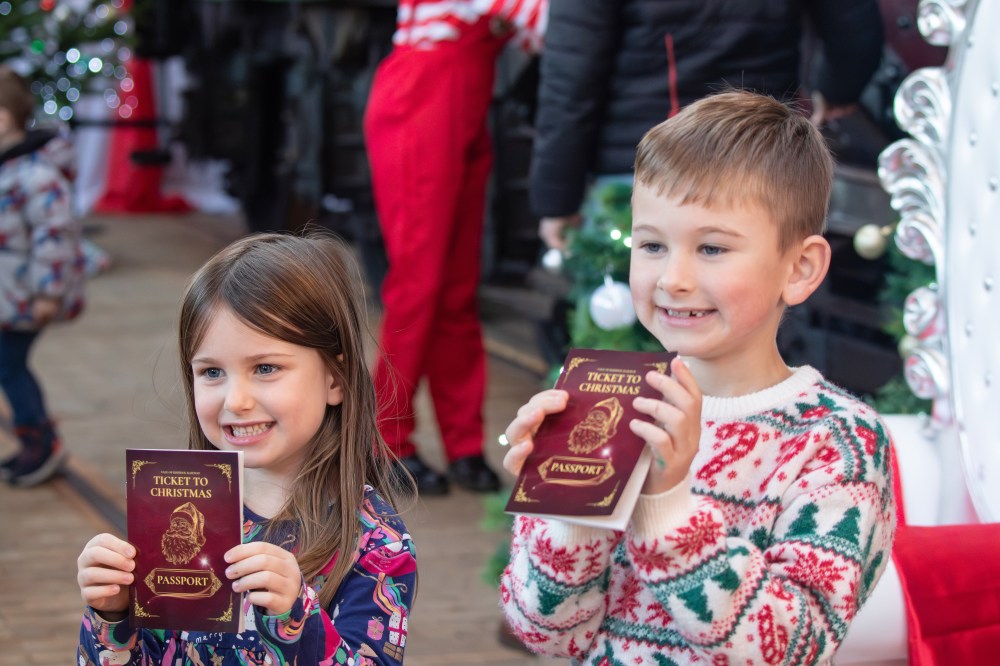 Two children hold 'Ticket to Christmas' booklets, smiling, standing indoors with festive decorations.