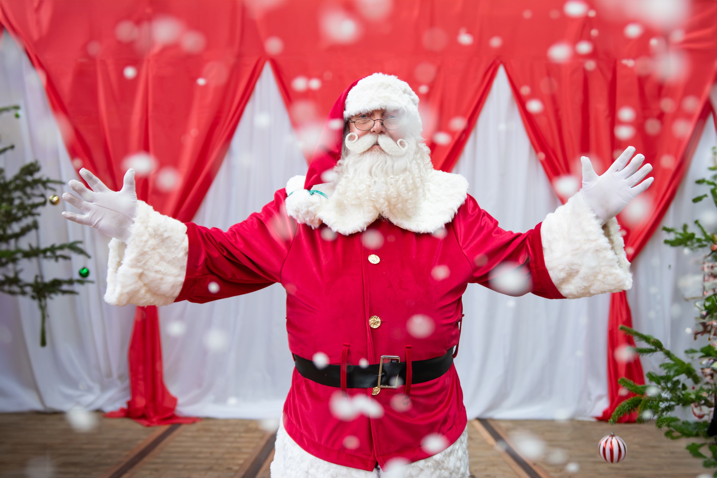 Santa Claus with arms open, surrounded by falling snow, red curtains in background.