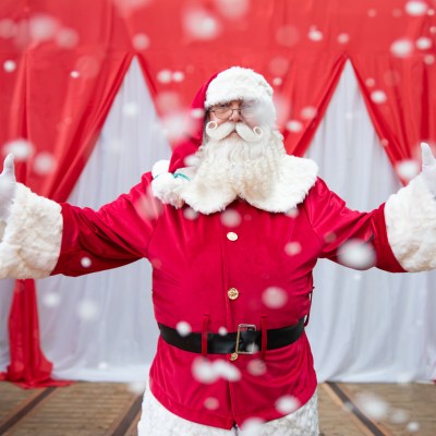 Santa Claus with arms open, surrounded by falling snow, red curtains in background.