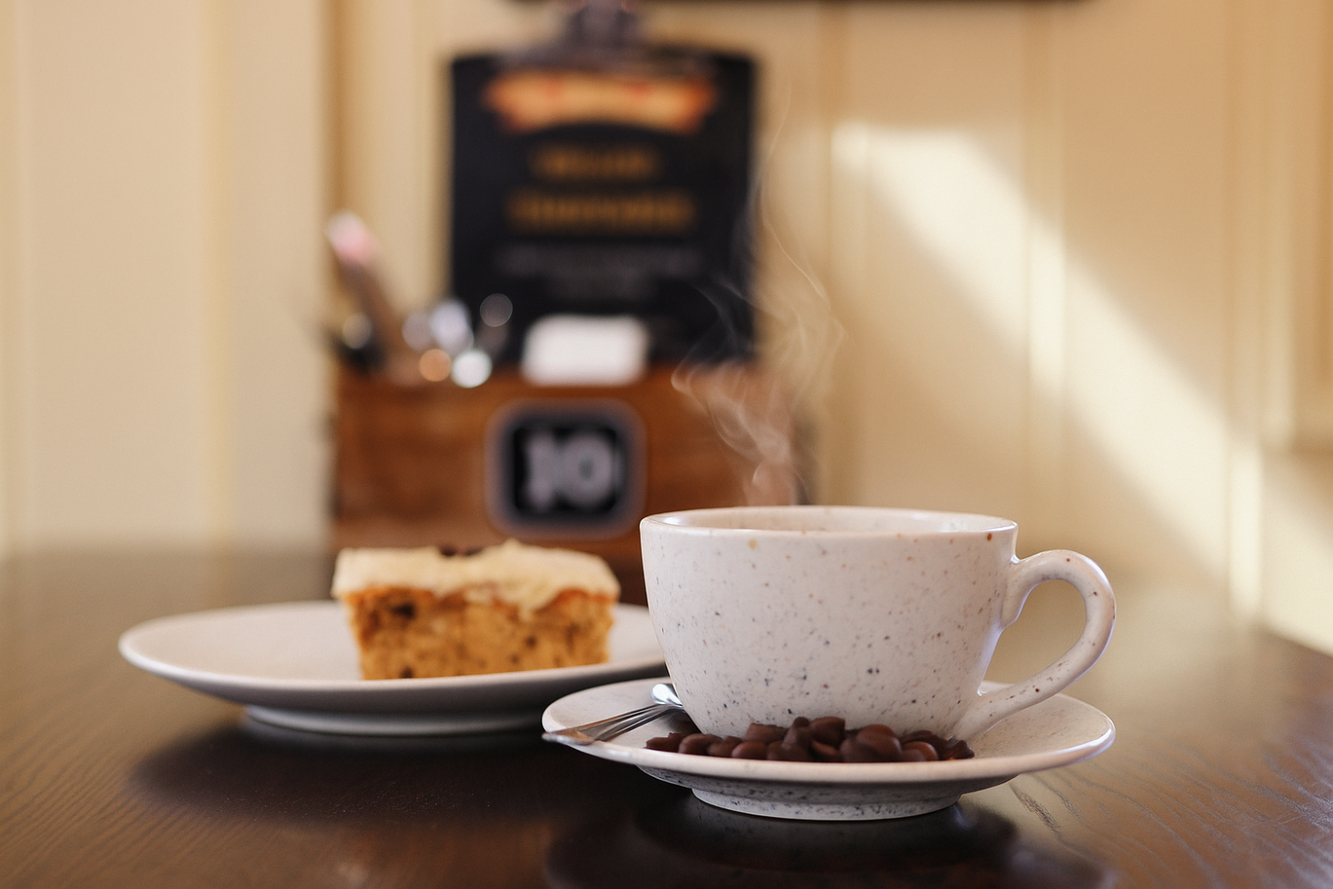 Steaming coffee cup on saucer with coffee beans, cake in the background.