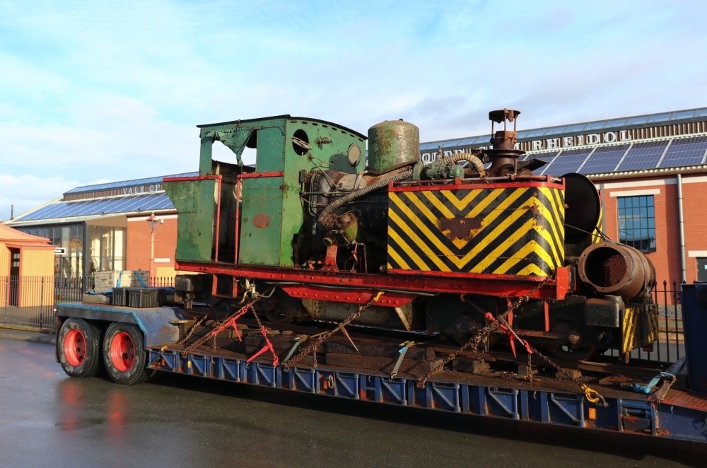 Old green locomotive on a flatbed trailer in front of a red brick building.