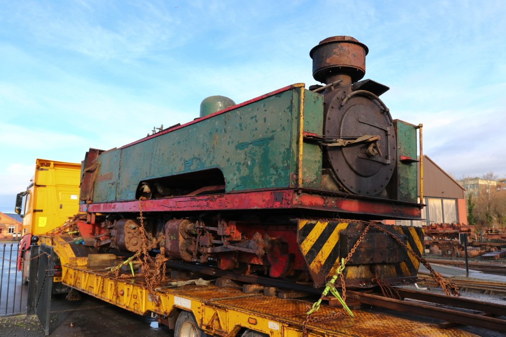 A vintage steam locomotive on a flatbed trailer under a blue sky.