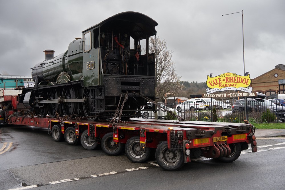 Steam locomotive on a trailer in front of a railway sign under cloudy skies.