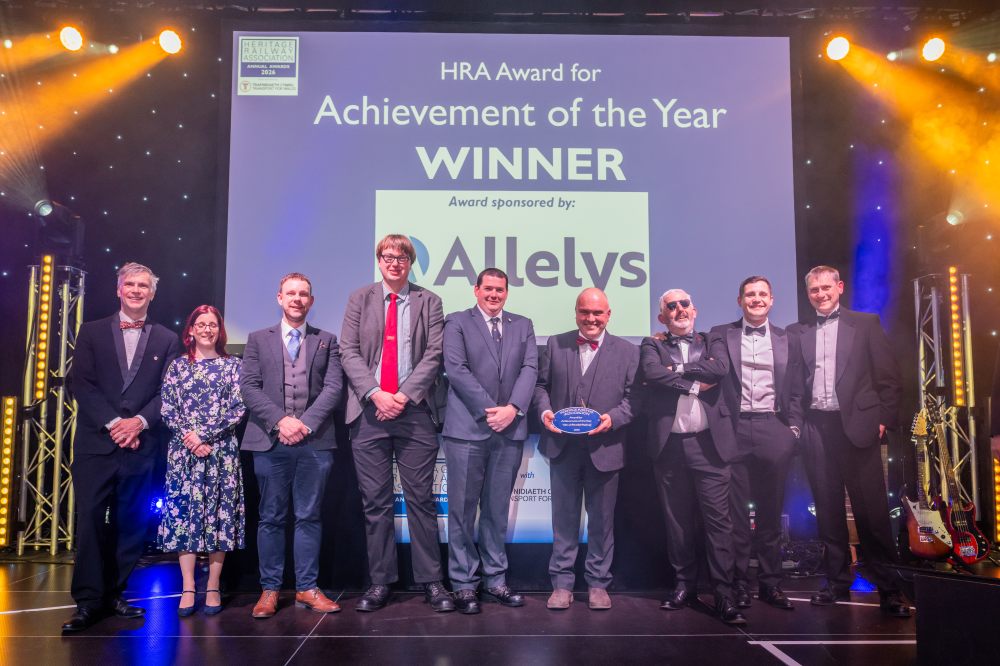 Group photo of award winners on stage holding a plaque, with a large screen displaying 'Achievement of the Year'.