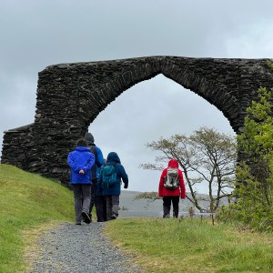 Four people walk towards a large stone arch on a cloudy day.
