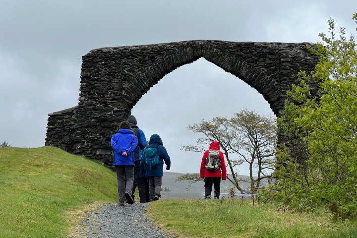 Four people walk towards a large stone arch on a cloudy day.