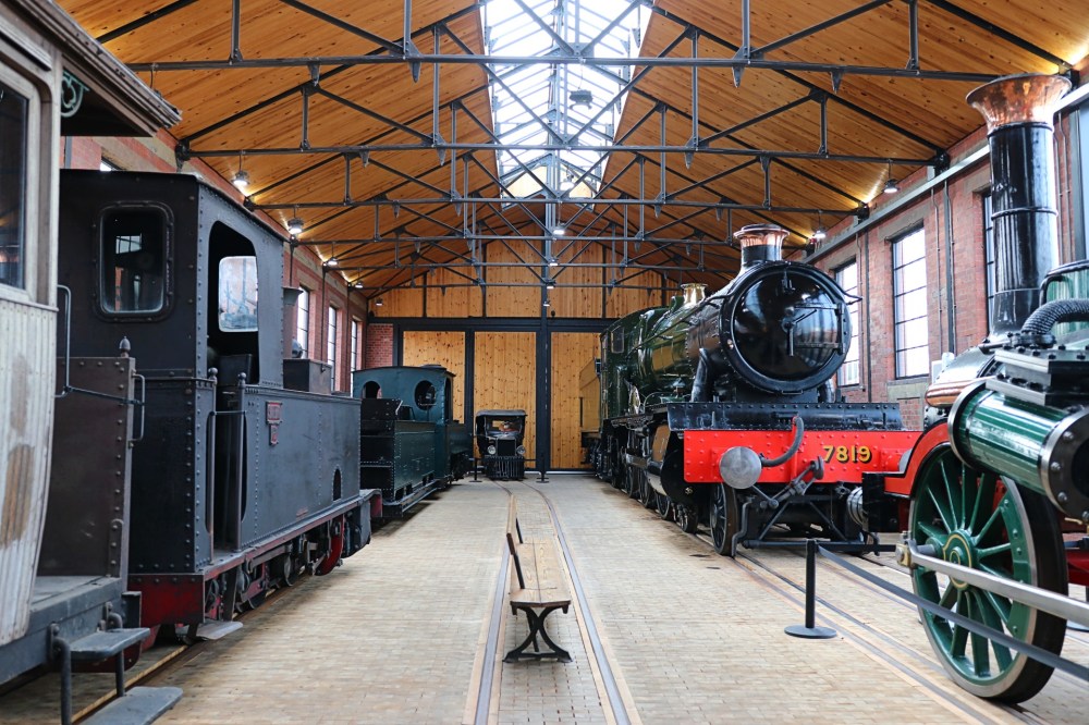 Interior of a train museum with vintage steam locomotives on display and wooden ceiling.