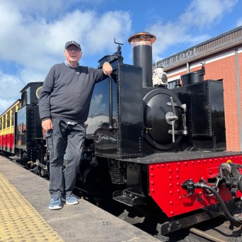 Man standing beside a vintage steam train on a sunny day.
