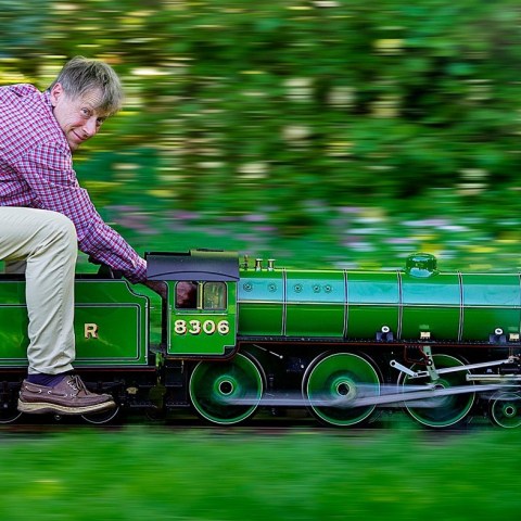 Person rides miniature green train swiftly in garden setting.