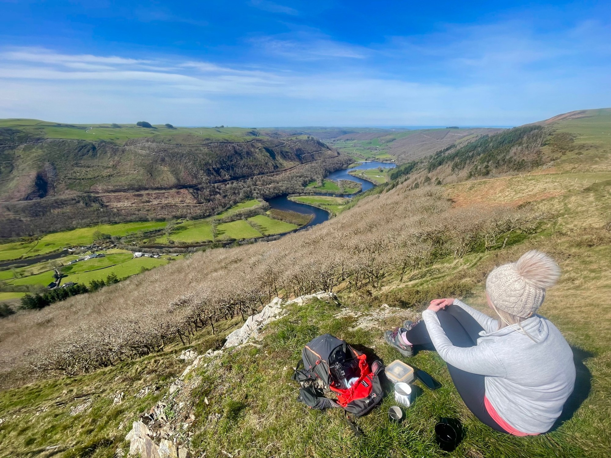 Person sitting on a hill overlooking a valley and river with a backpack and cup nearby.