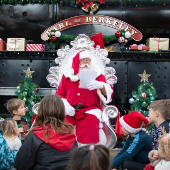 Santa with children seated around him in front of a decorated train.