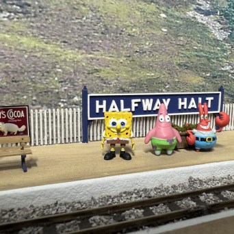 Toy figures stand on a model train platform labeled 'Halfway Halt'.