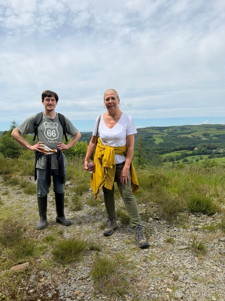Two hikers stand on a trail with a scenic hilly landscape in the background.