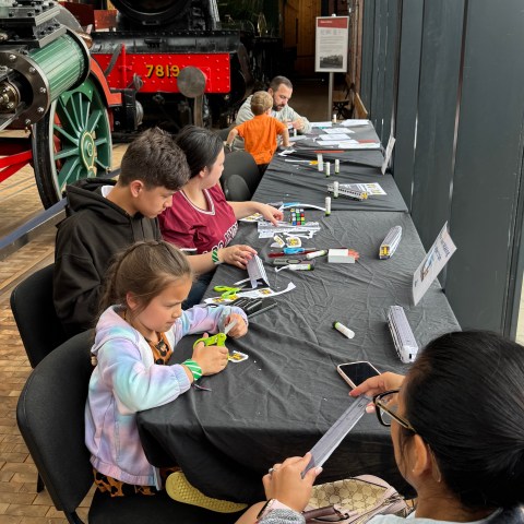 Children sit at a table doing crafts in a train museum, with a steam engine in the background.
