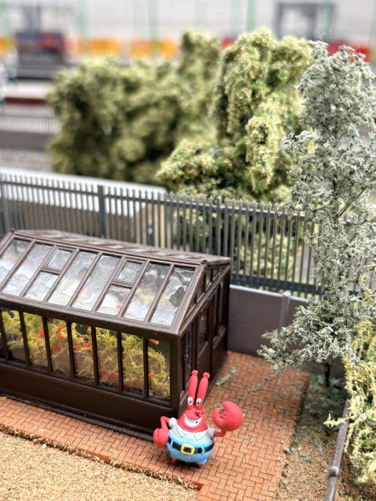 Toy figure near greenhouse, surrounded by greenery and pathway.