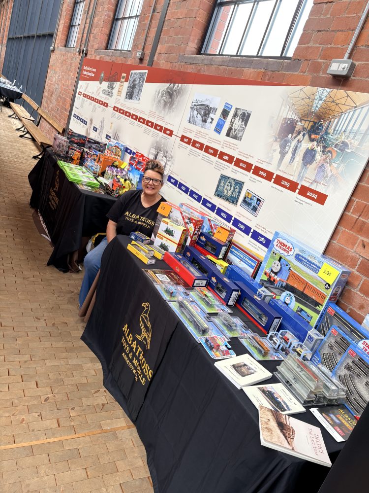 Person with glasses sits at a vendor table with model trains and books on display.