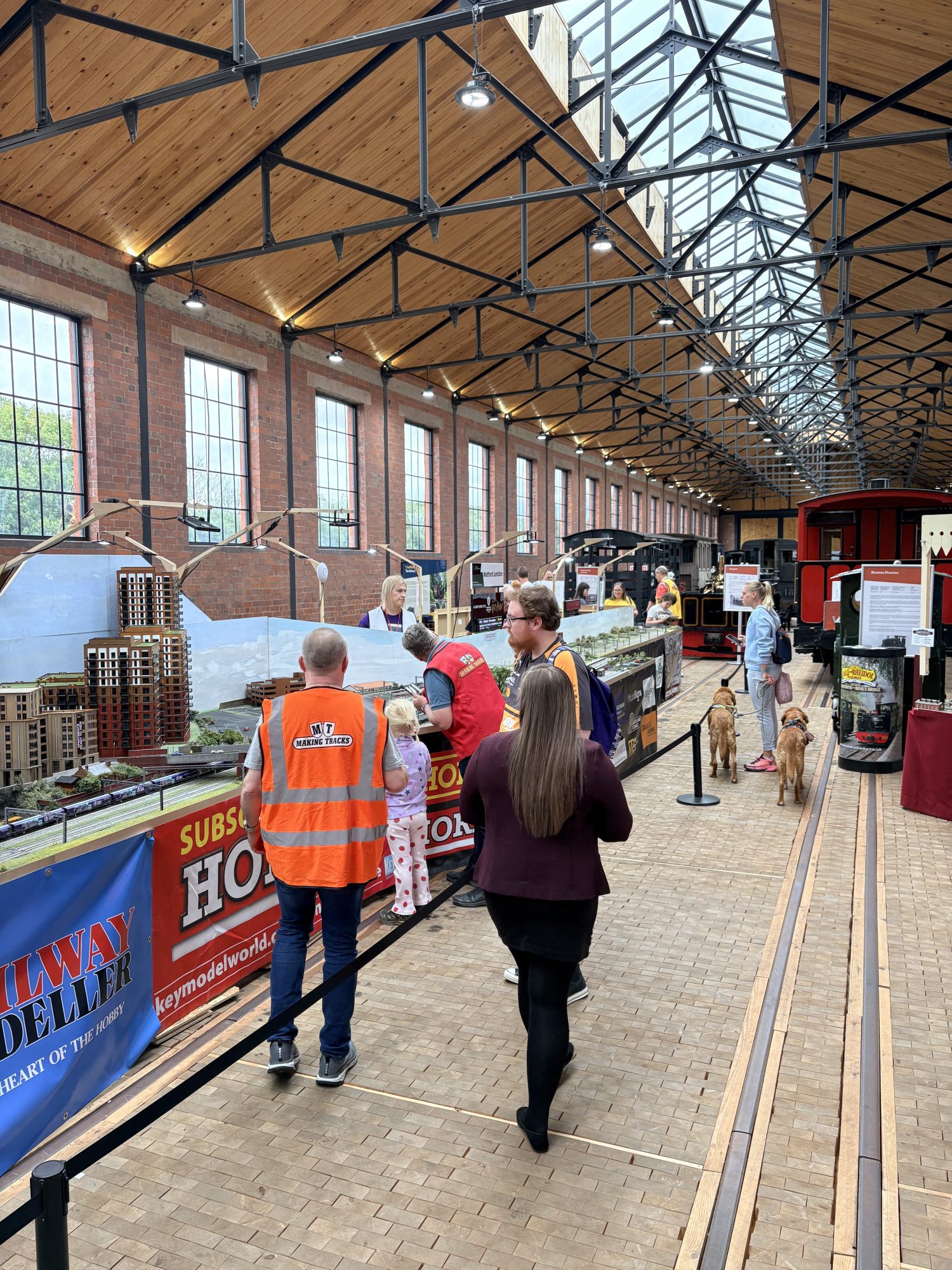 People view model train exhibit in large hall with high ceiling and brick walls.