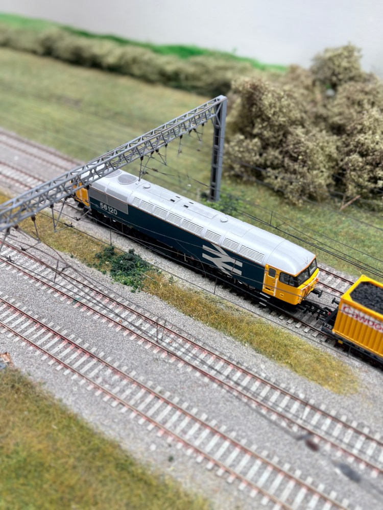 Model train on tracks with overhead wires and greenery.
