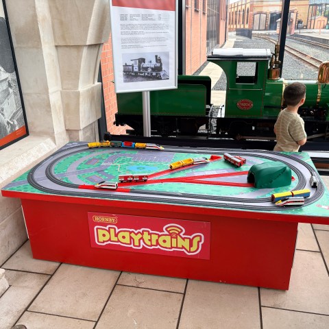 Child at a model train display on a red table labeled 'Playtrains' with a green locomotive nearby.