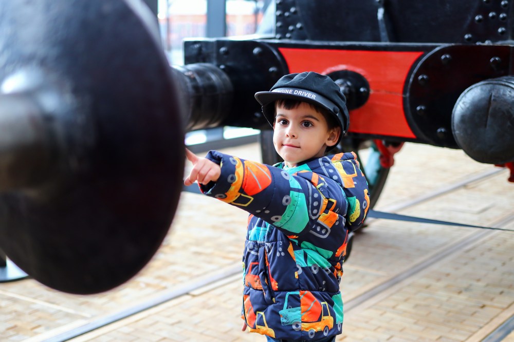 Child in colorful jacket pointing, standing near large black machinery.